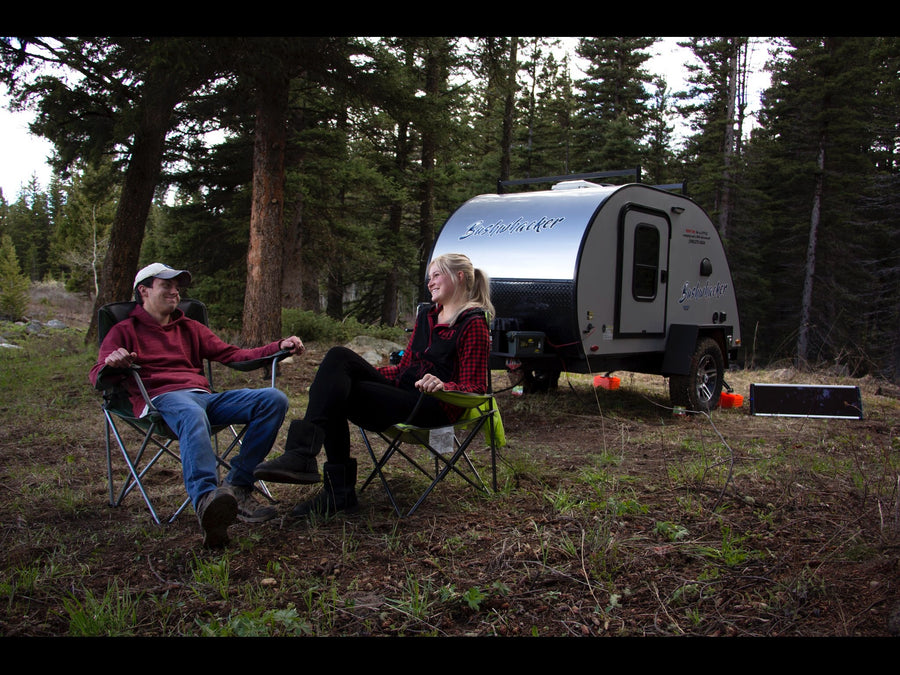 Two people sit in chairs by a Bushwhacker RV rental at a wooded campsite, enjoying the outdoors.