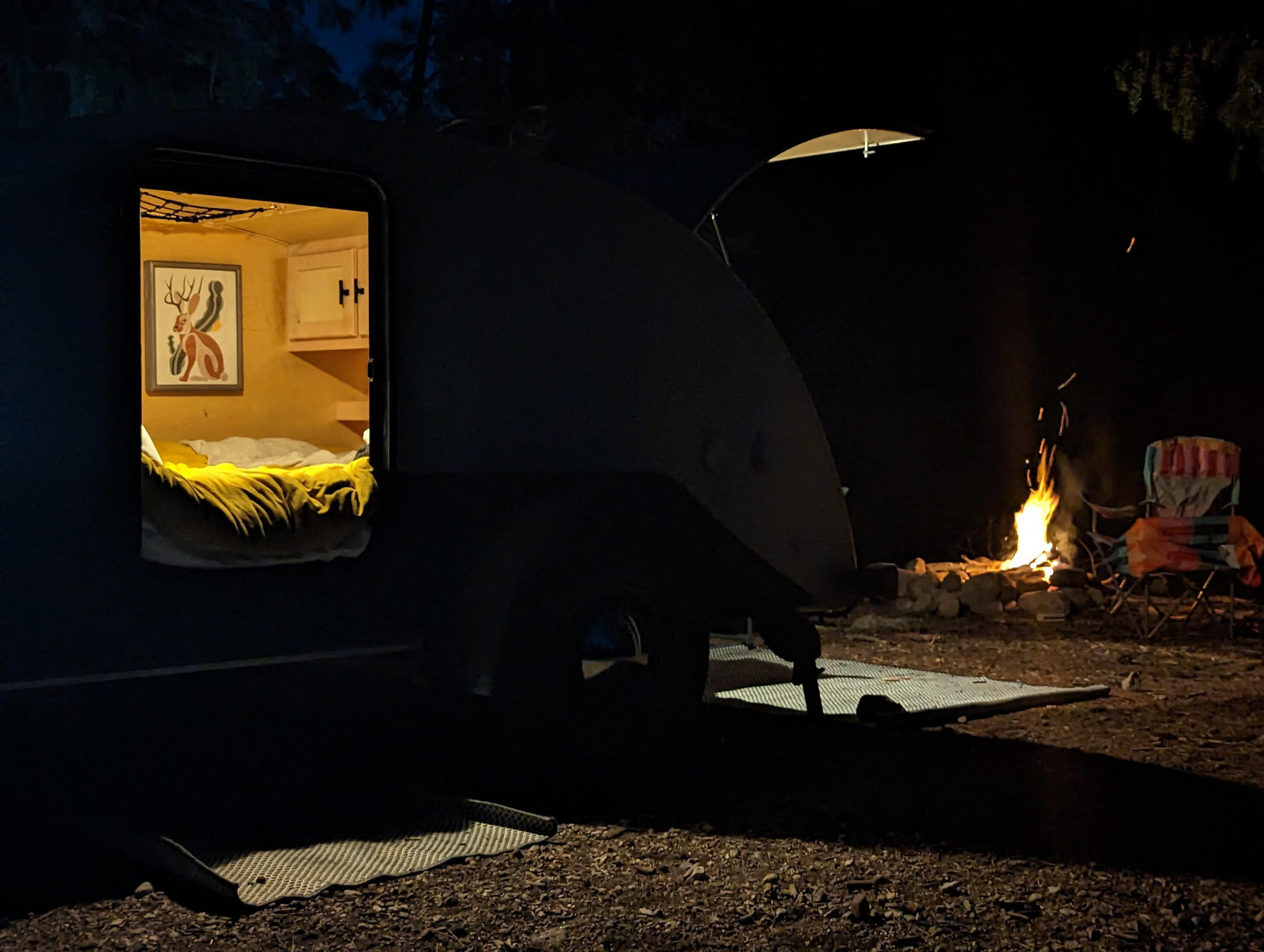 and a person by the trailer.|Experience camping! A stylish grey teardrop RV rental parked on a forest campsite with mountain views.|Inside a cozy RV rental: a bed with yellow sheets and white bedding