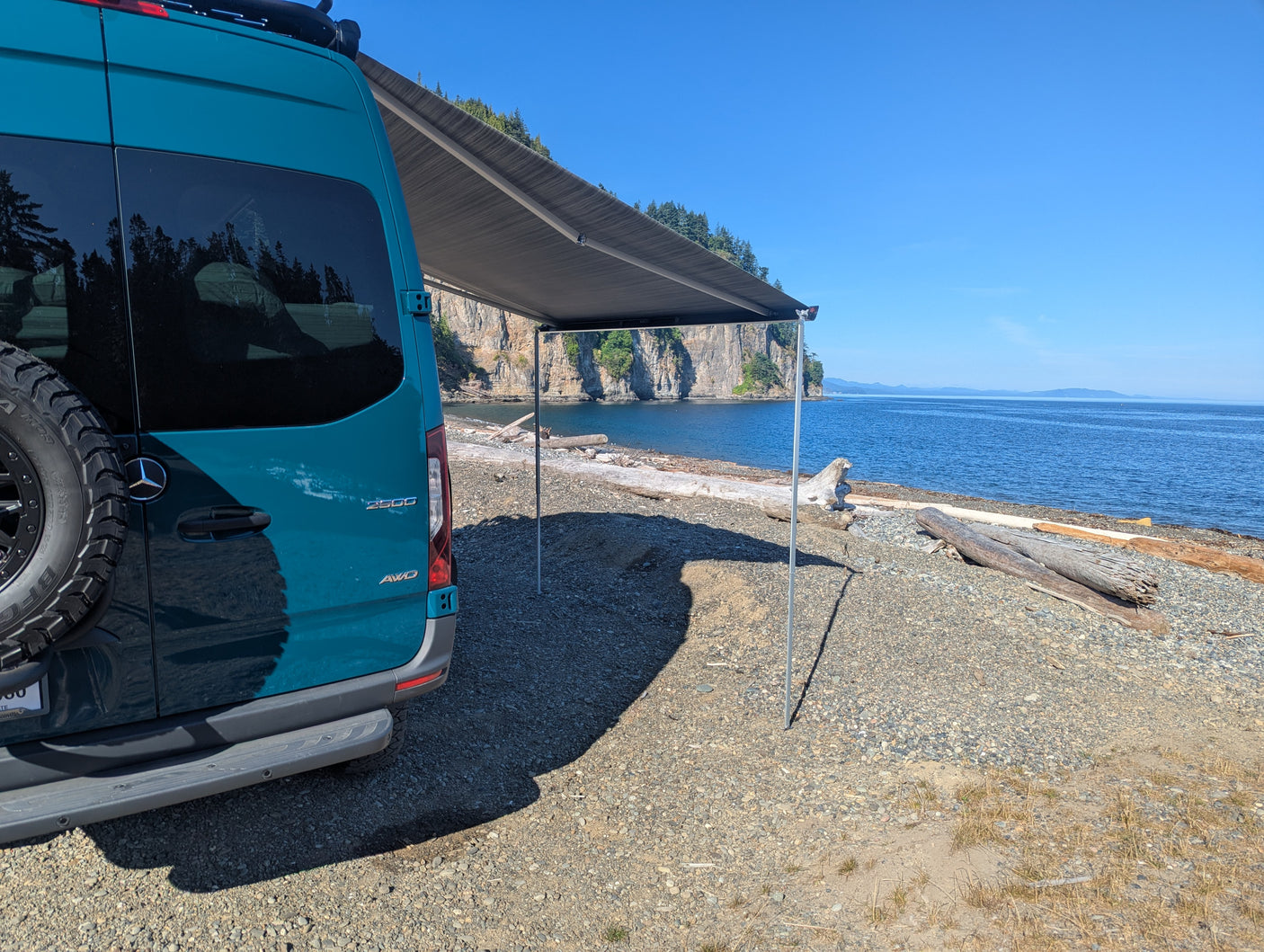 Teal RV rental with awning extended on a rocky beach by blue water & cliffs. Perfect for adventure!
