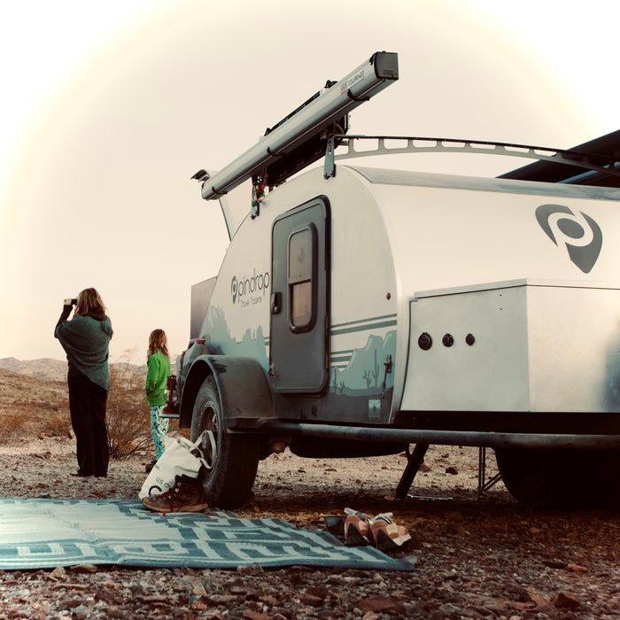 An RV rental teardrop trailer by Pindrop, set in a desert landscape, with two people looking out at the scenic views.