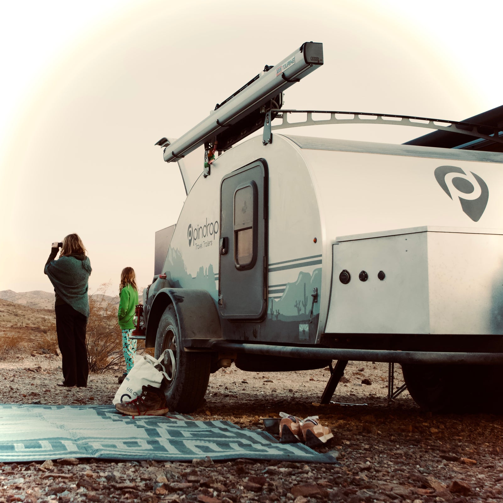 An RV rental teardrop trailer by Pindrop, set in a desert landscape, with two people looking out at the scenic views.