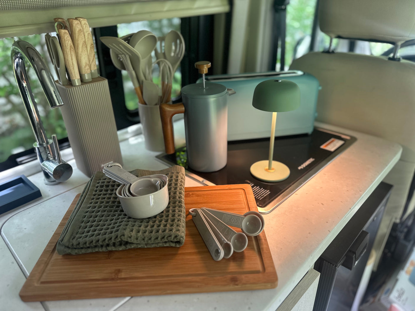 Compact RV rental kitchen counter with knives, cooking utensils, French press, toaster, measuring cups, and a portable lamp.