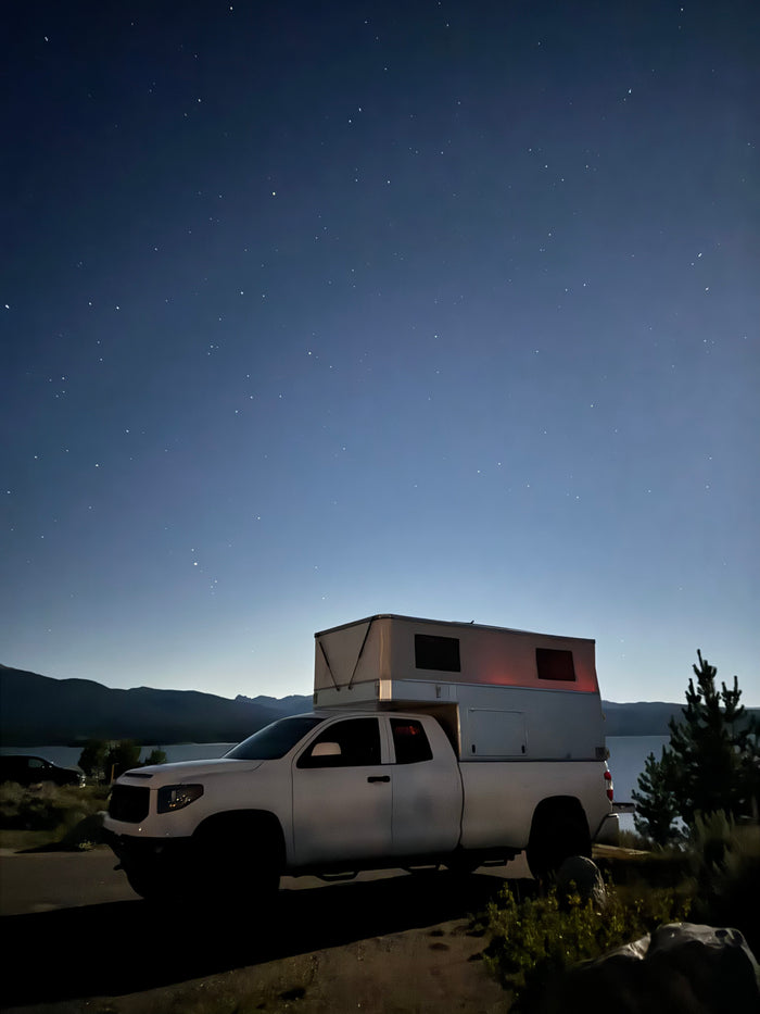 This white RV rental truck camper is parked by a lake under a clear, starry night sky, perfect for outdoor adventures.
