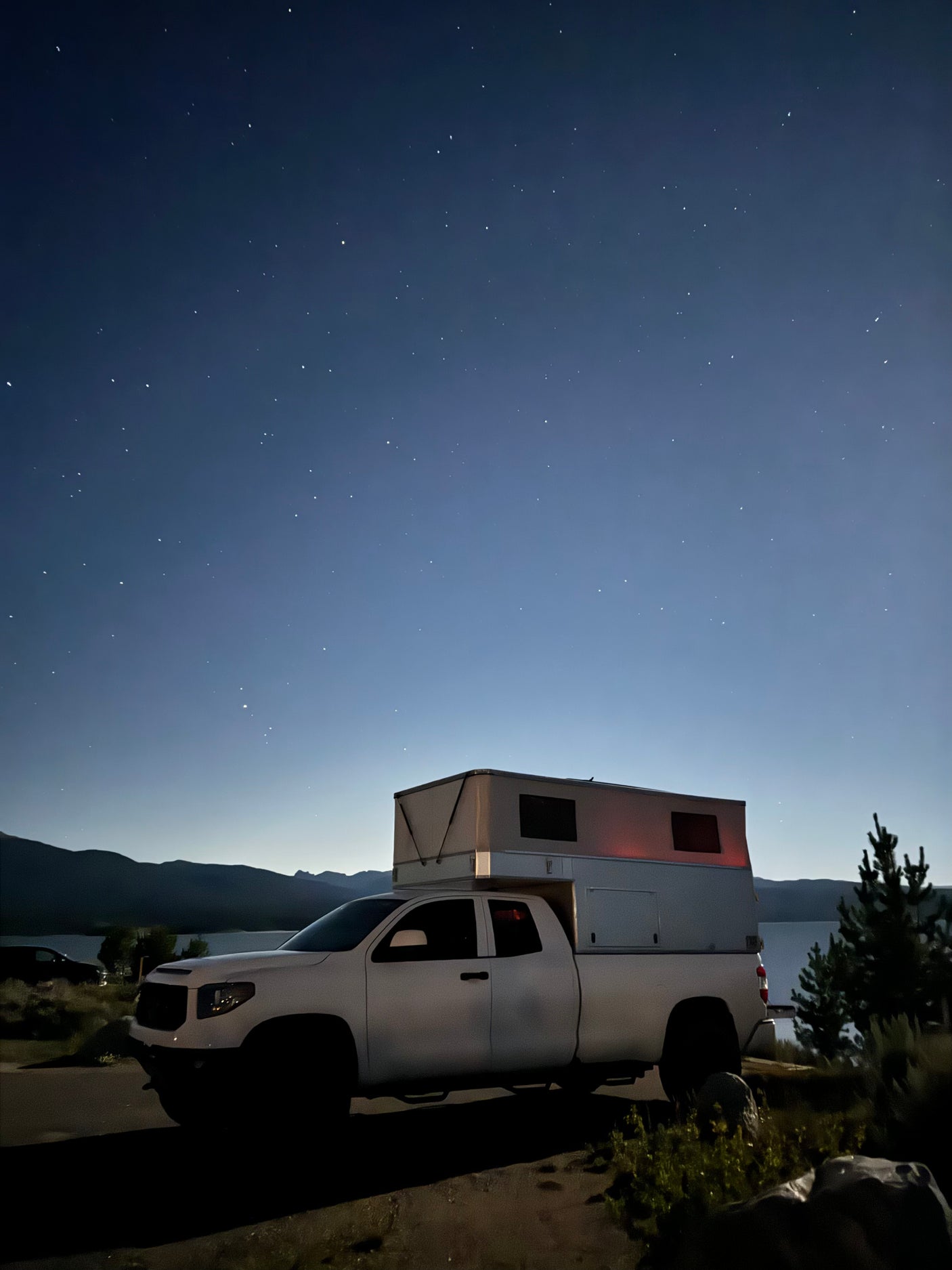 This white RV rental truck camper is parked by a lake under a clear, starry night sky, perfect for outdoor adventures.