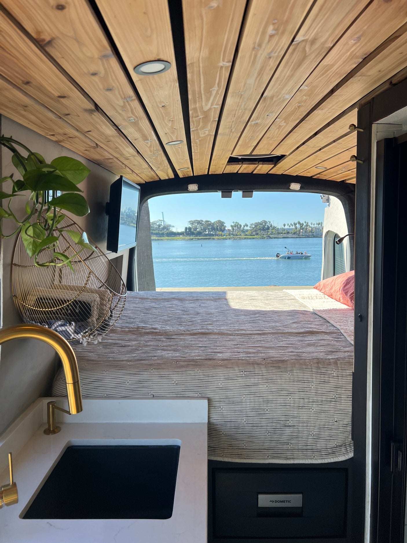 RV rental: Interior shot from the front showing a bed, kitchen counter with gold faucet, and a Dometic fridge, with a stunning water view from the rear.