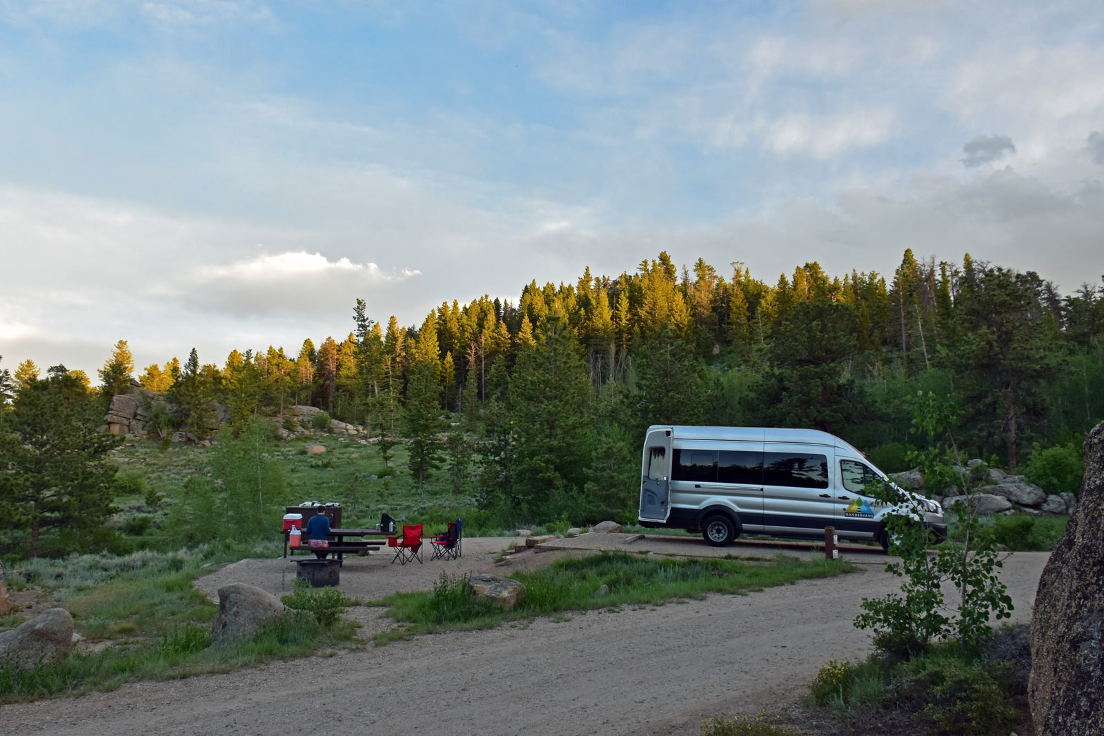 A silver RV rental van is parked at a quiet campsite in a forested mountain landscape, with a person at a picnic table.