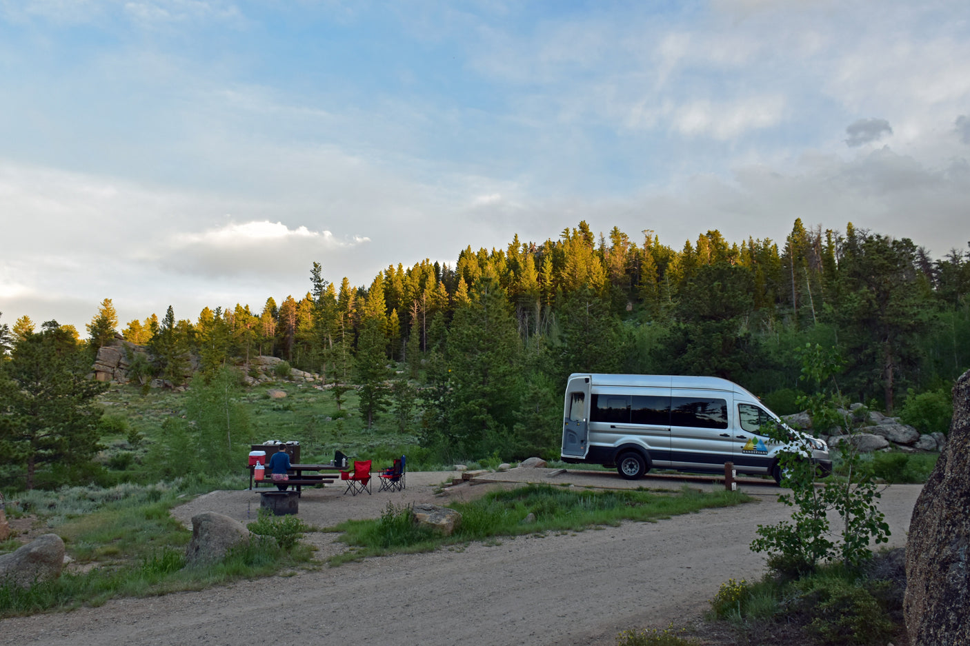 A silver RV rental van is parked at a quiet campsite in a forested mountain landscape, with a person at a picnic table.
