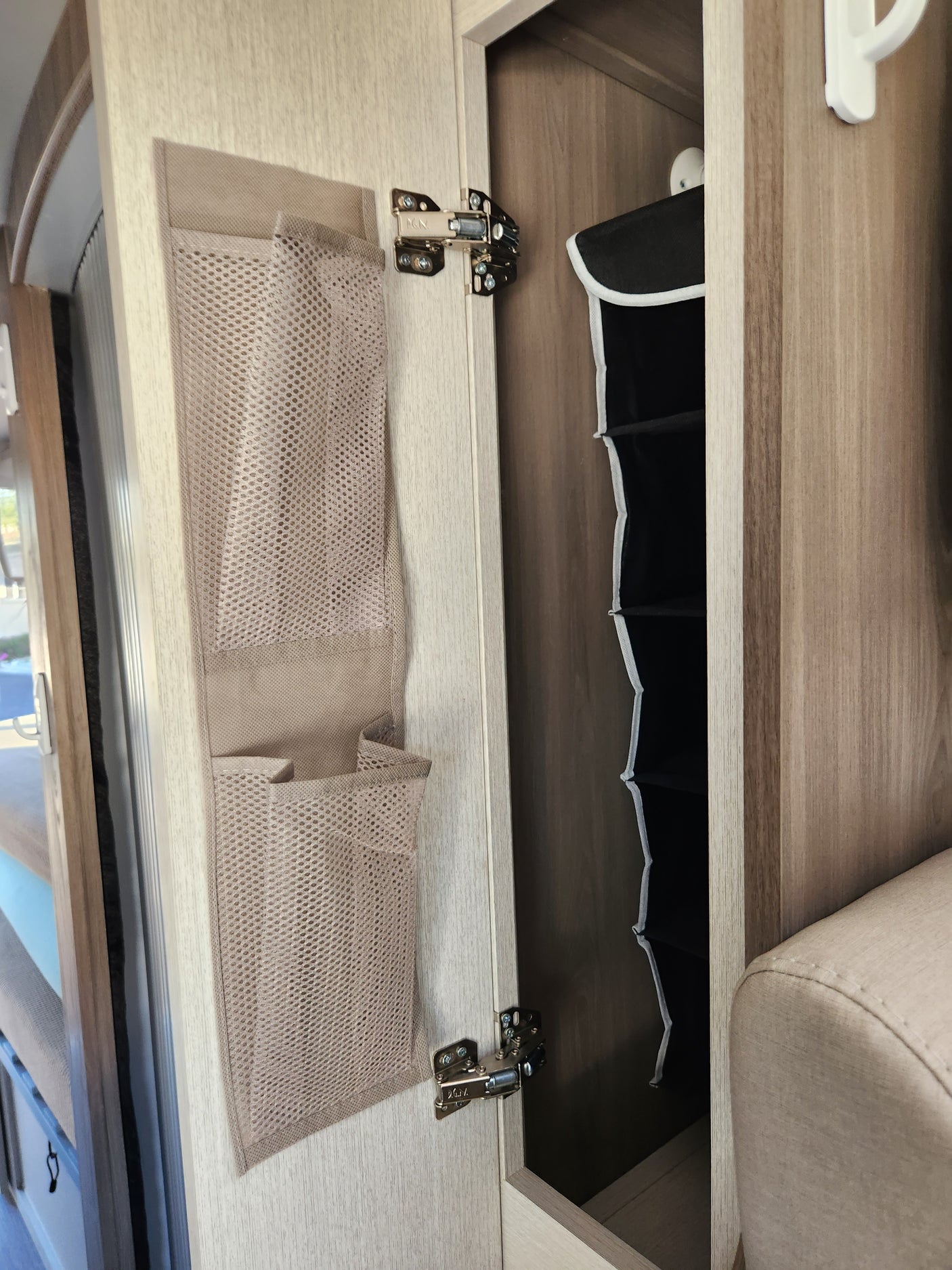 Inside an RV rental: an open overhead cabinet reveals a black foldable organizer. The door has a beige mesh pocket system, above a window.