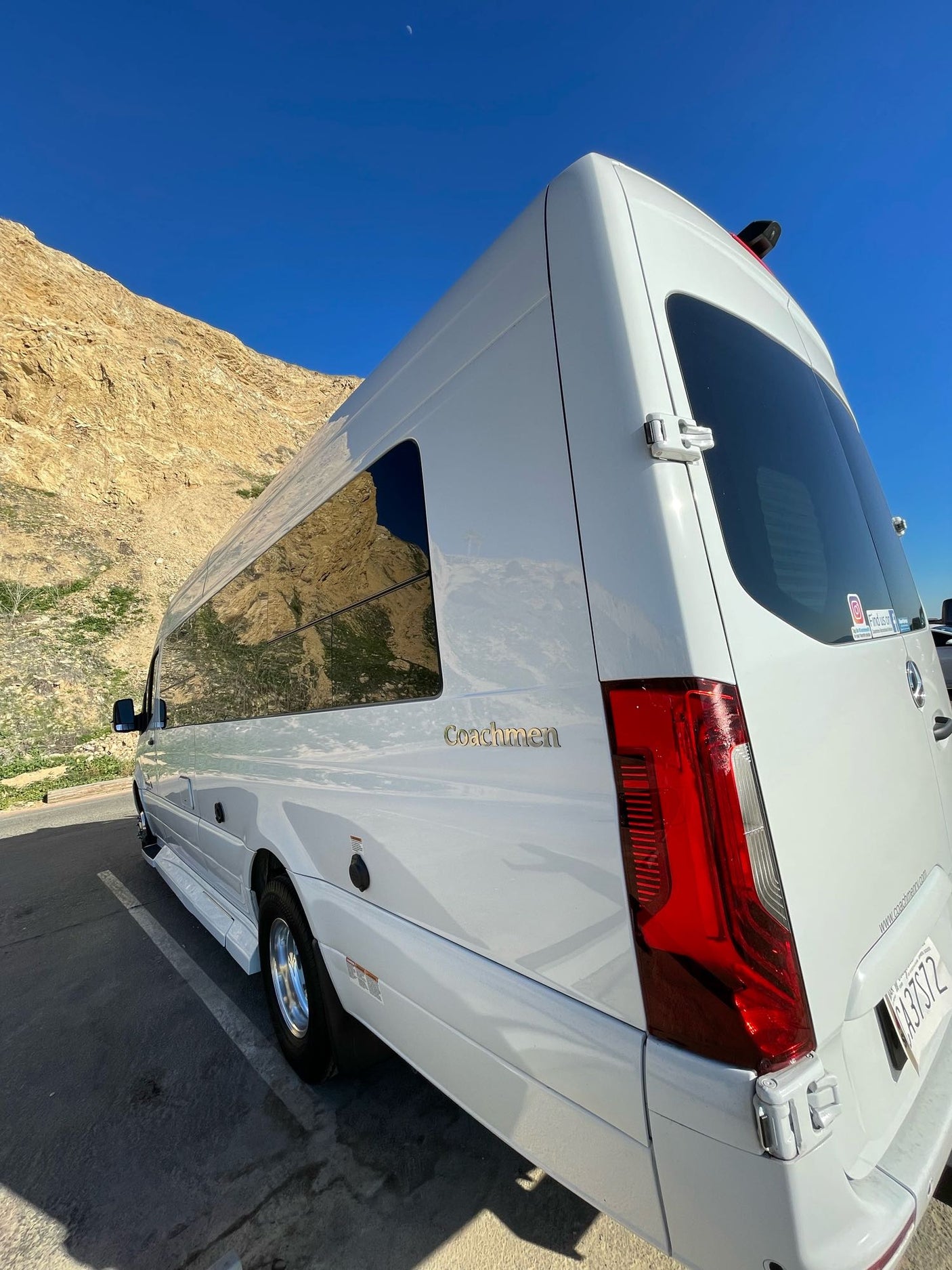 White Coachmen RV rental parked by sunny mountains under a blue sky with a crescent moon. Perfect for a road trip!