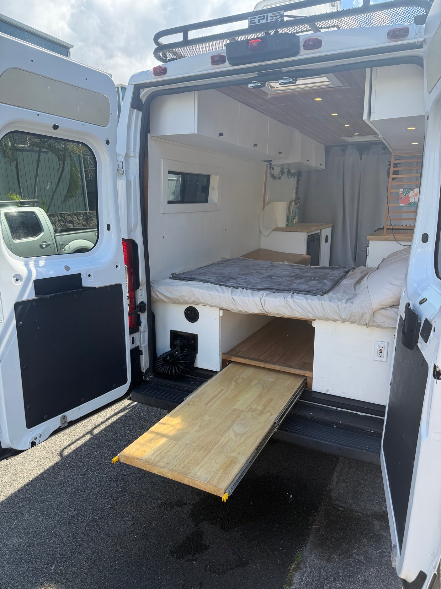 Rear view of an RV rental: a white camper van with open doors, showing a bed, kitchen, storage, and a pull-out wooden tray.