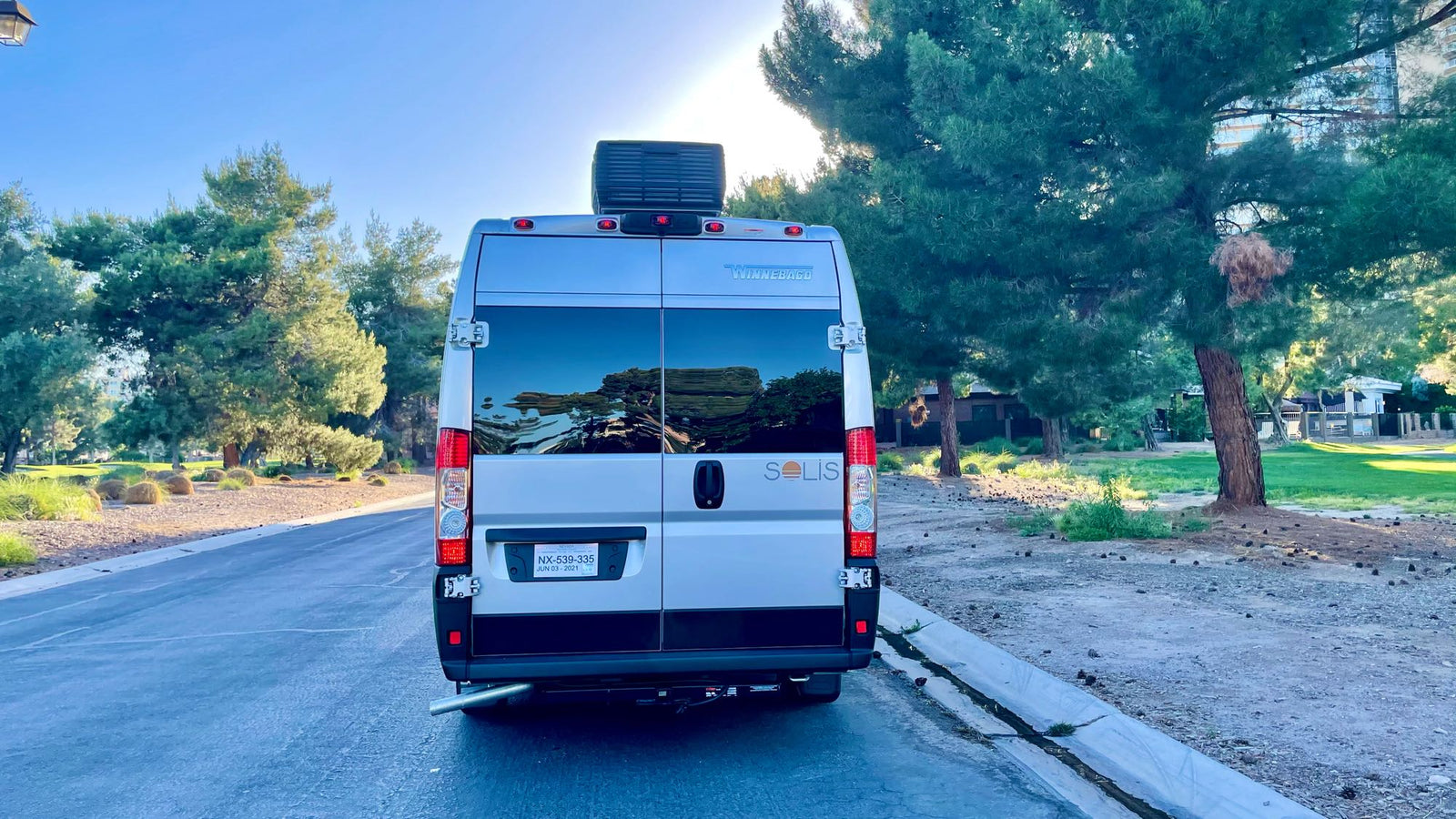 Silver Winnebago Solis RV rental parked on a sunny road, its rear reflecting trees. Ready for a great camper van trip!