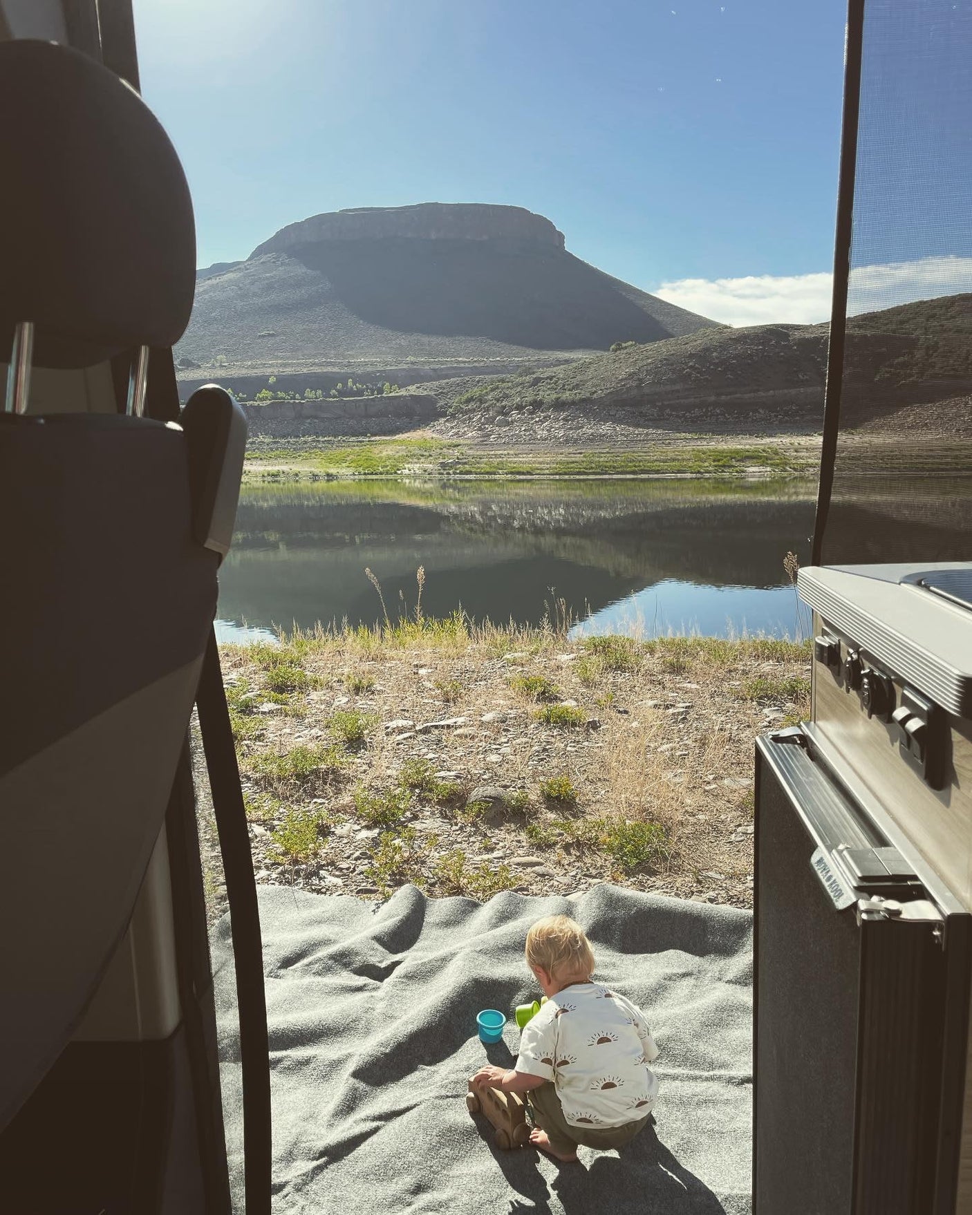 A child sits on a blanket by an rv rental, facing a tranquil lake with mountains under a sunny sky. Perfect family camp day.
