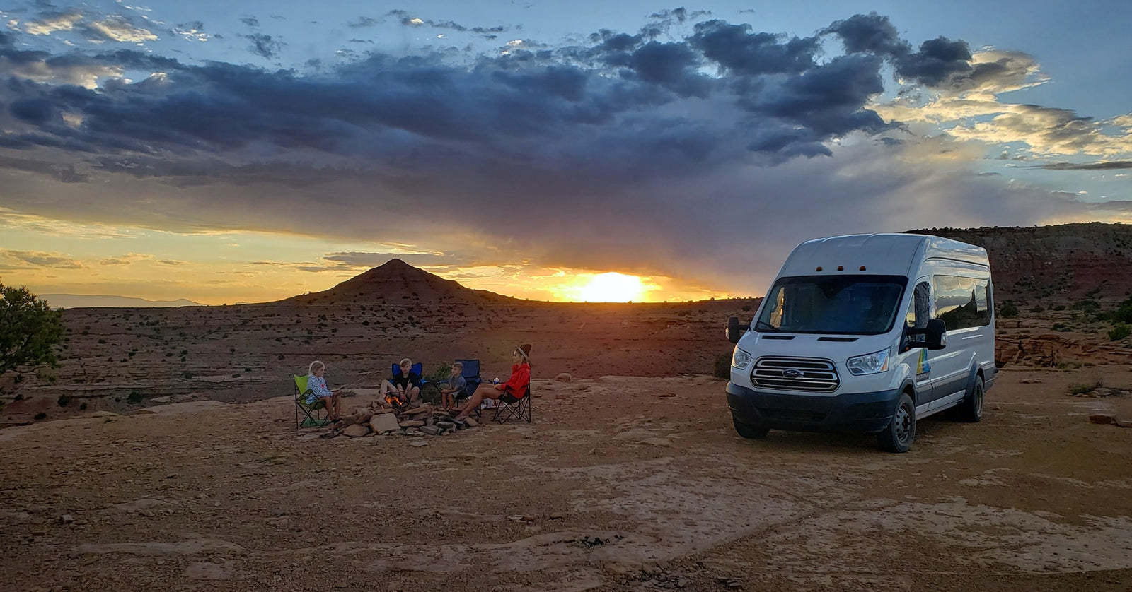 An RV rental parked at sunset, as a family gathers around a warm campfire in a vast desert landscape.