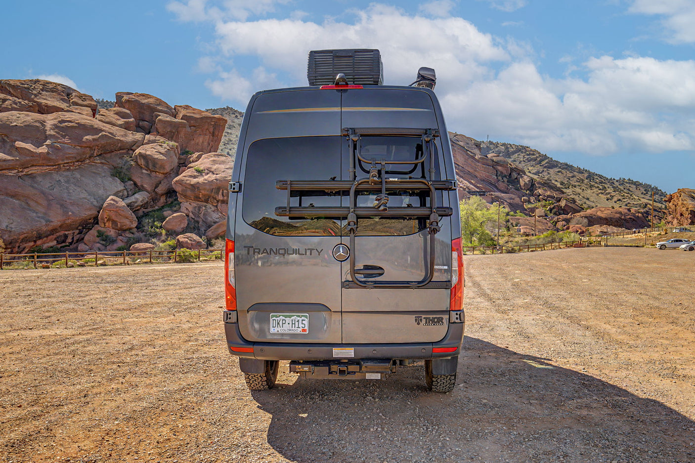 Grey Tranquility RV rental with a bike rack, parked in a dirt lot against a backdrop of rocky mountains under a blue sky.