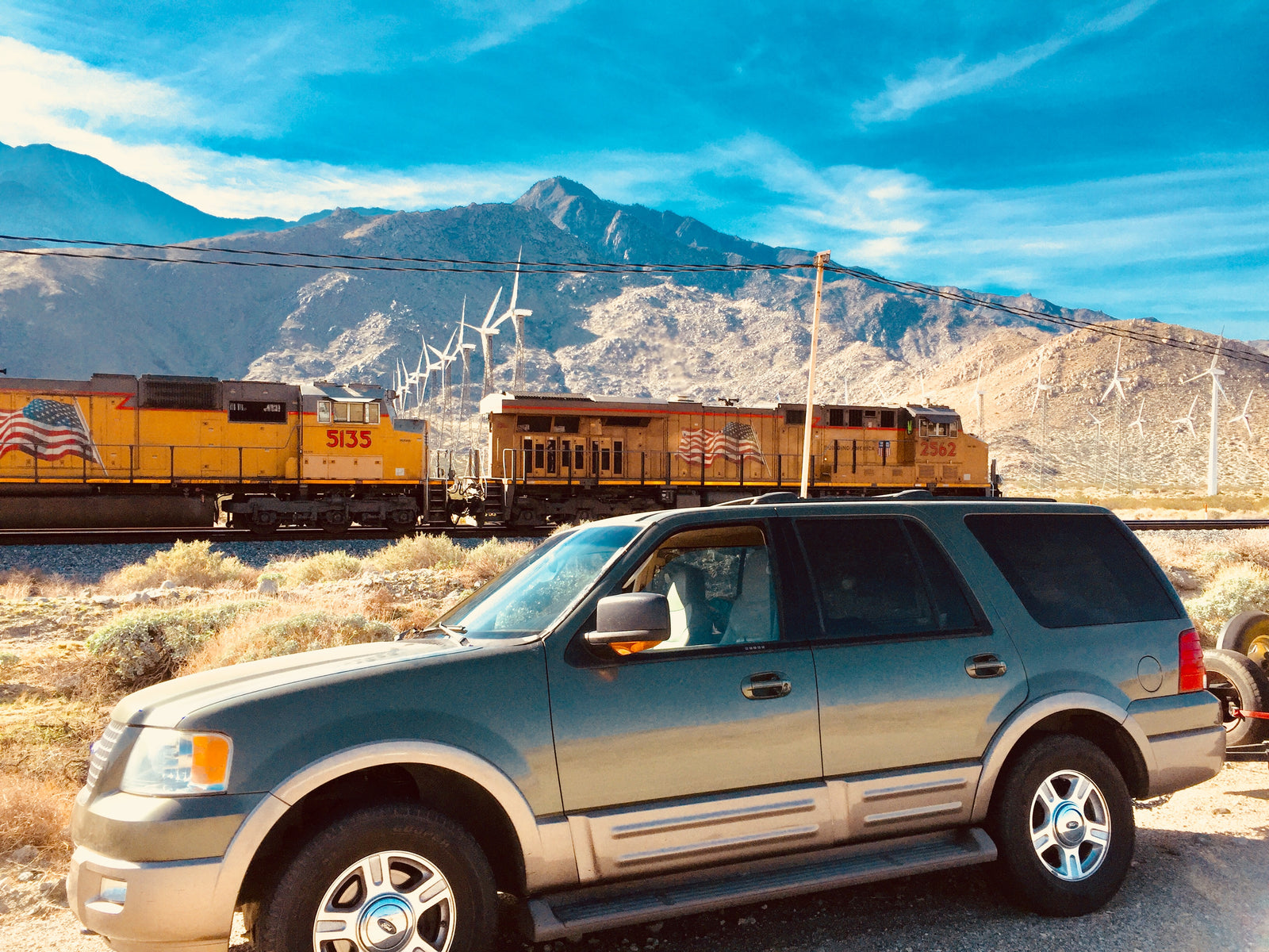 A gray SUV parked near train tracks, with a yellow train, wind turbines, and mountains under a blue sky, perfect for an rv rental adventure.