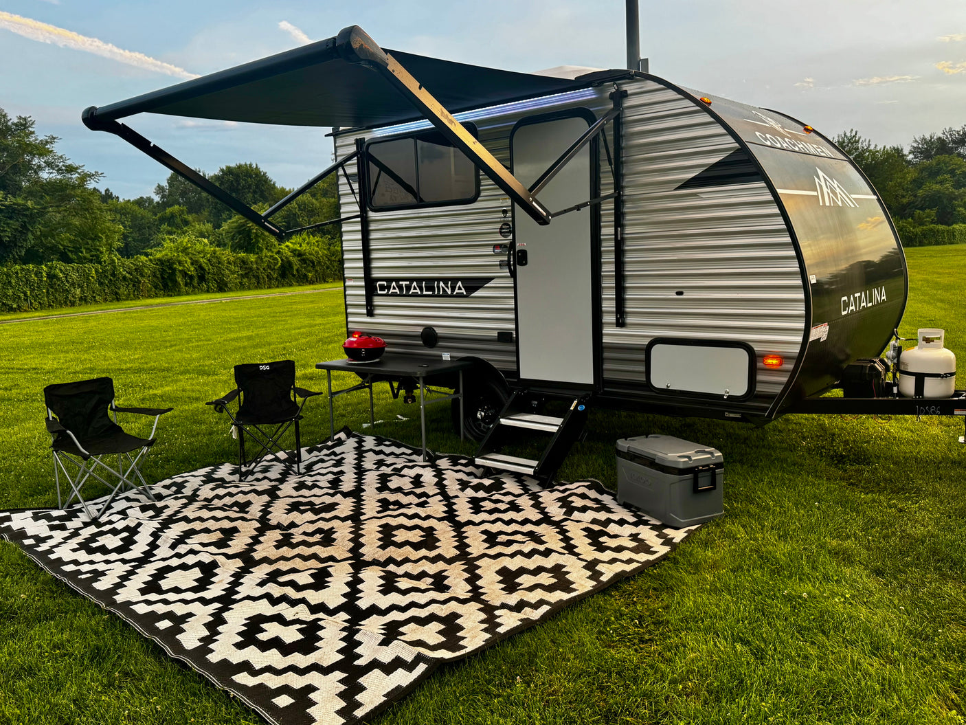 Experience the outdoors! An rv rental travel trailer with an extended awning, chairs, grill, and patterned rug on green grass.