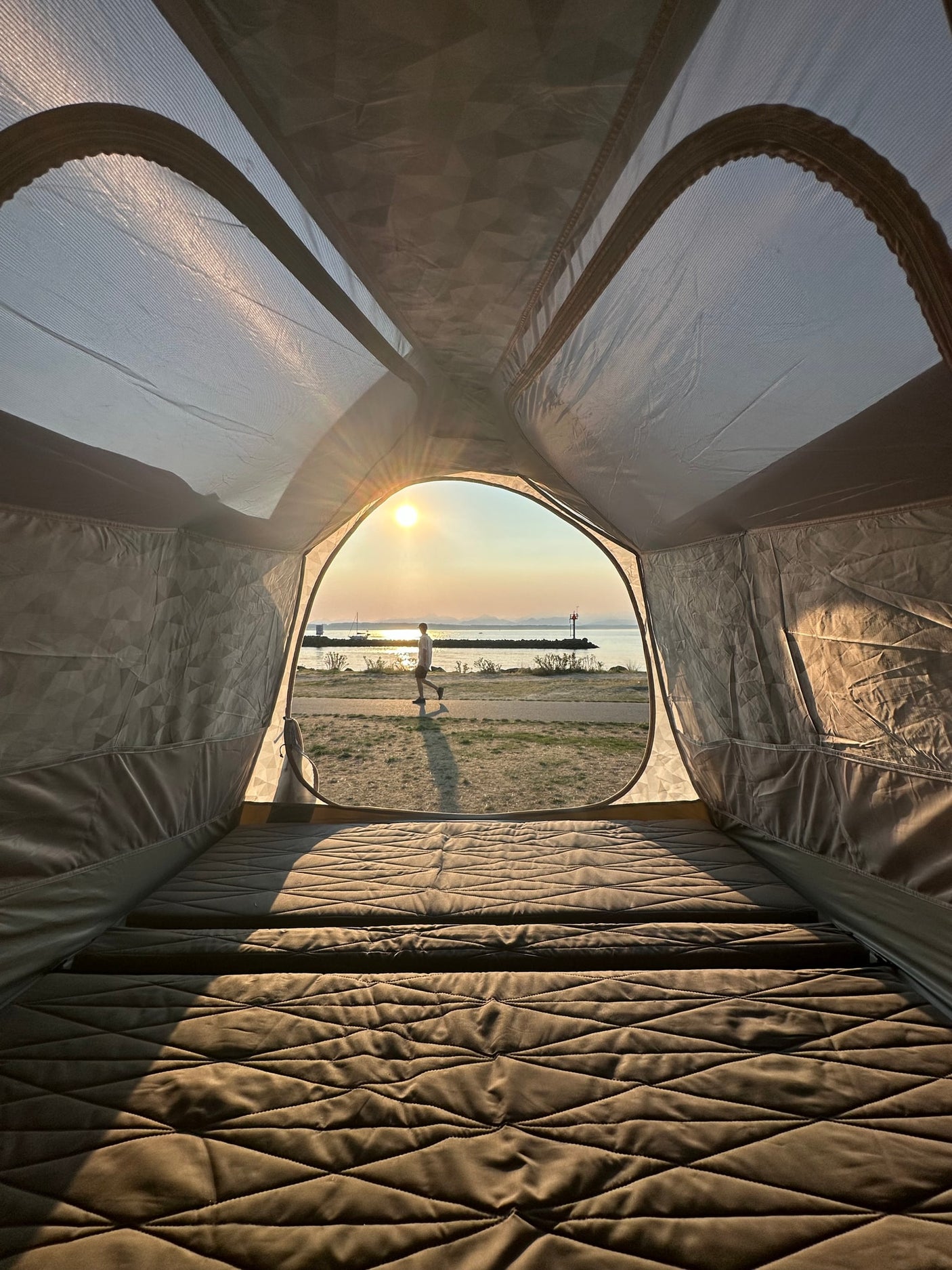 From an RV rental, a comfy quilted bed frames a golden sunset over water with a person walking outside.