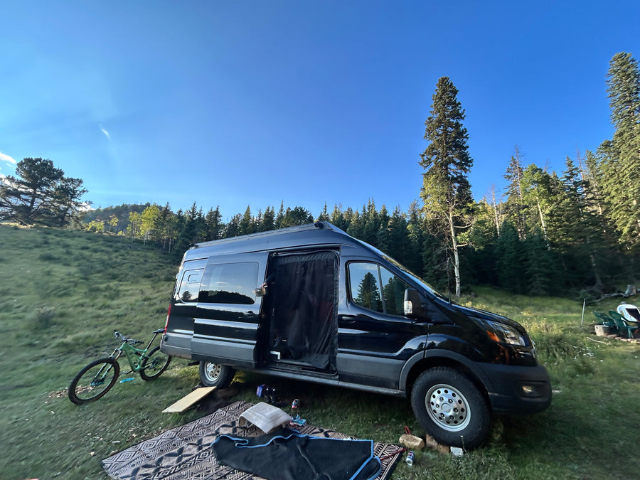 A black camper van RV rental is parked on a grassy slope near a forest, with a bike and outdoor setup for nature living.