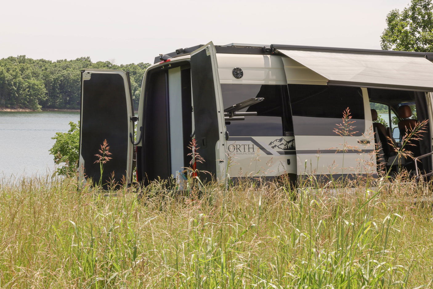 Experience outdoor freedom with this rv rental camper van, parked overlooking a tranquil lake with open doors and an extended awning.
