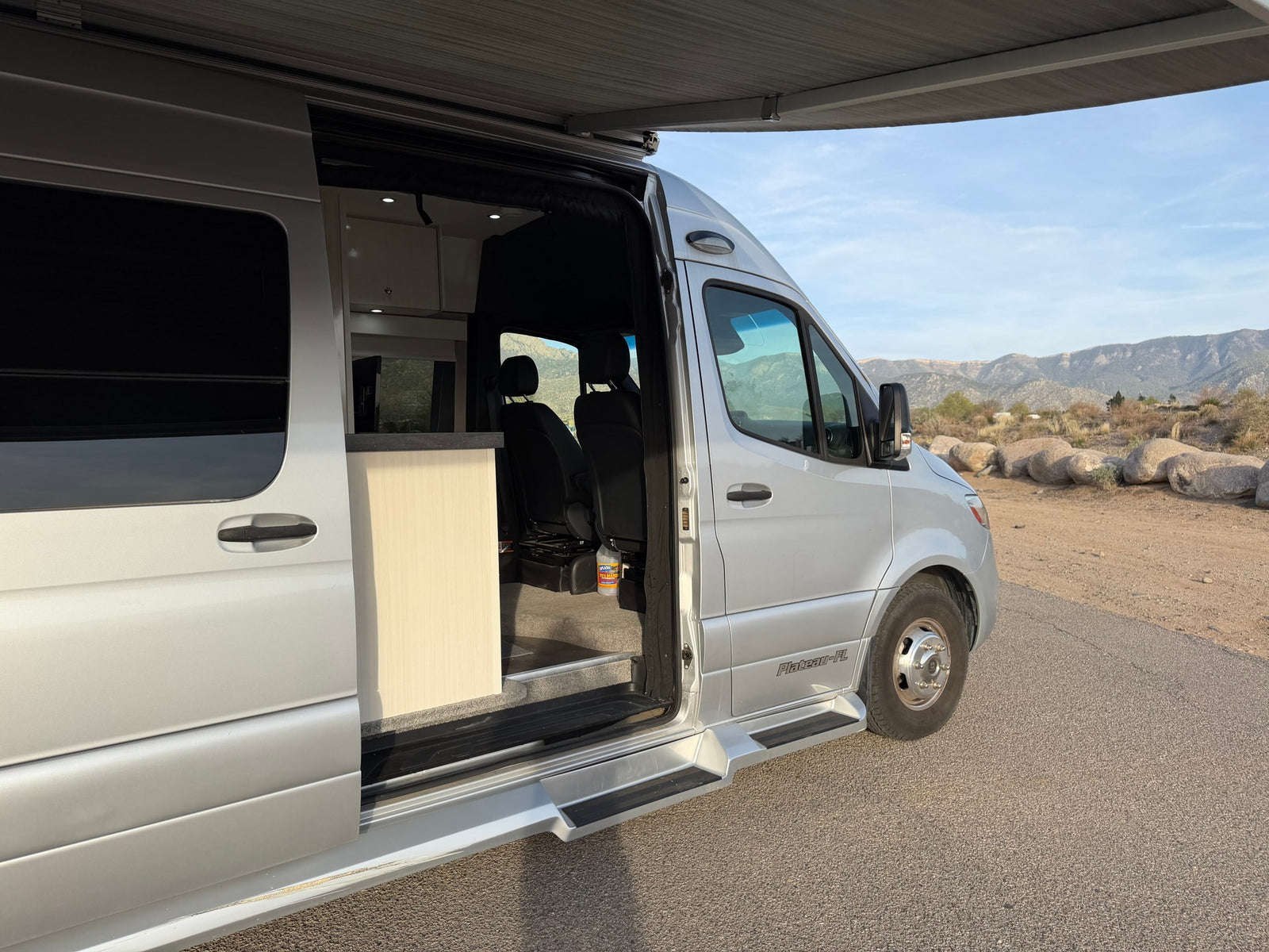 Explore in this silver RV rental camper van! Side door open, awning out, parked against a scenic mountain backdrop.
