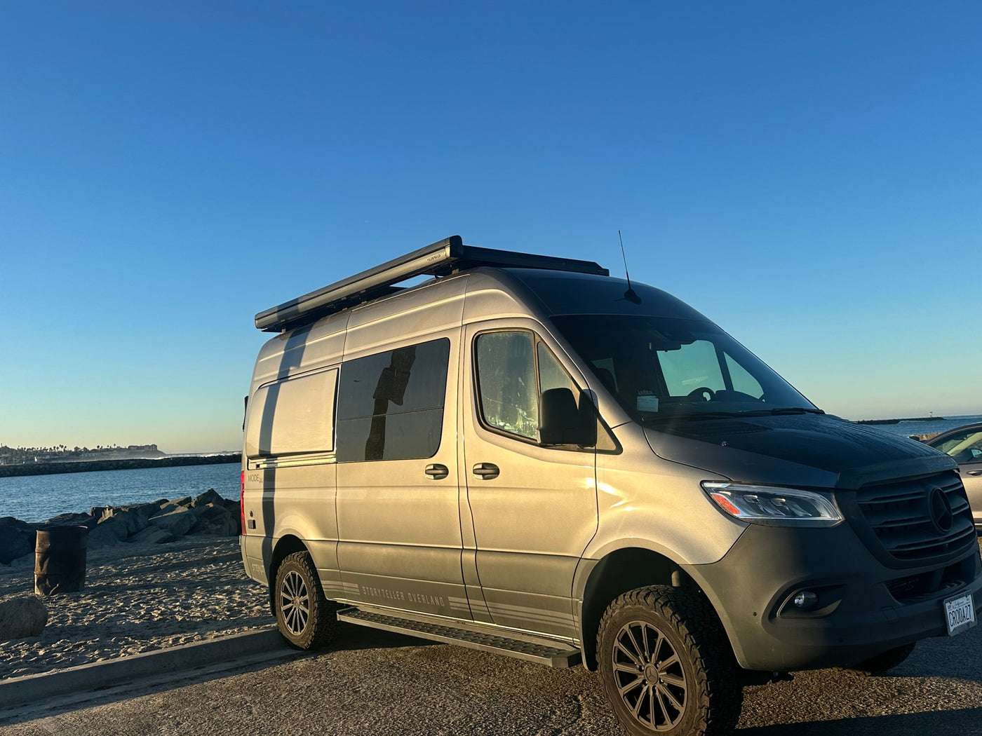 A grey Storyteller Overland Sprinter van RV rental is parked on a paved lot beside a sandy beach and ocean. — photo 1