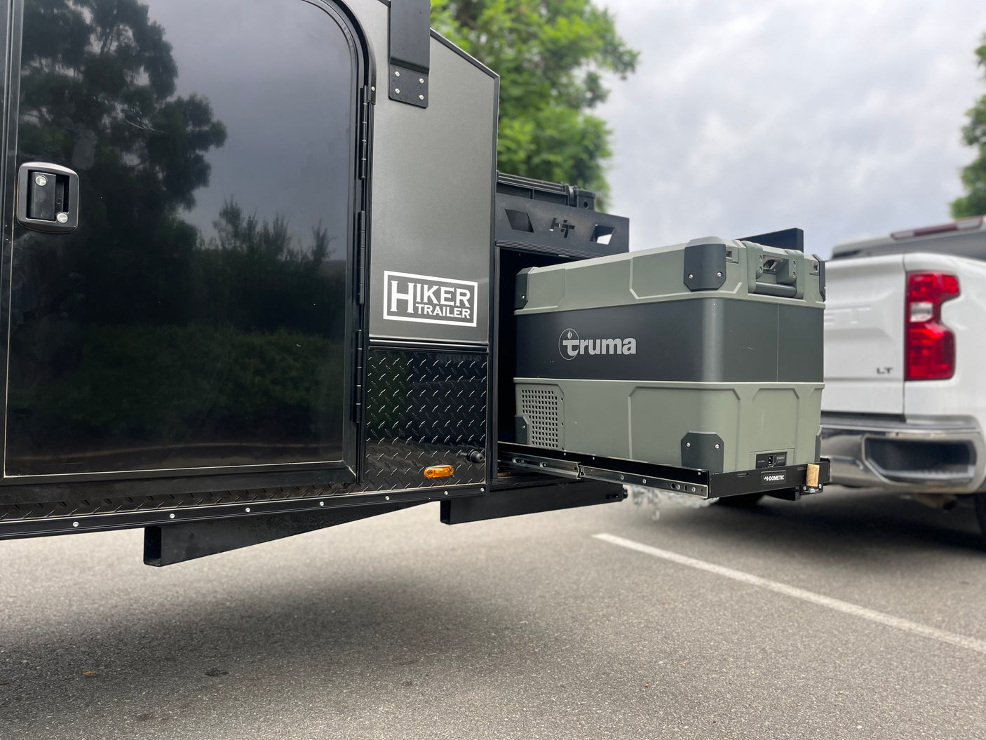 Hiker Trailer RV rental: A gray Truma portable fridge is pulled out on a Dometic slide from the side of the trailer. Ideal camping setup.