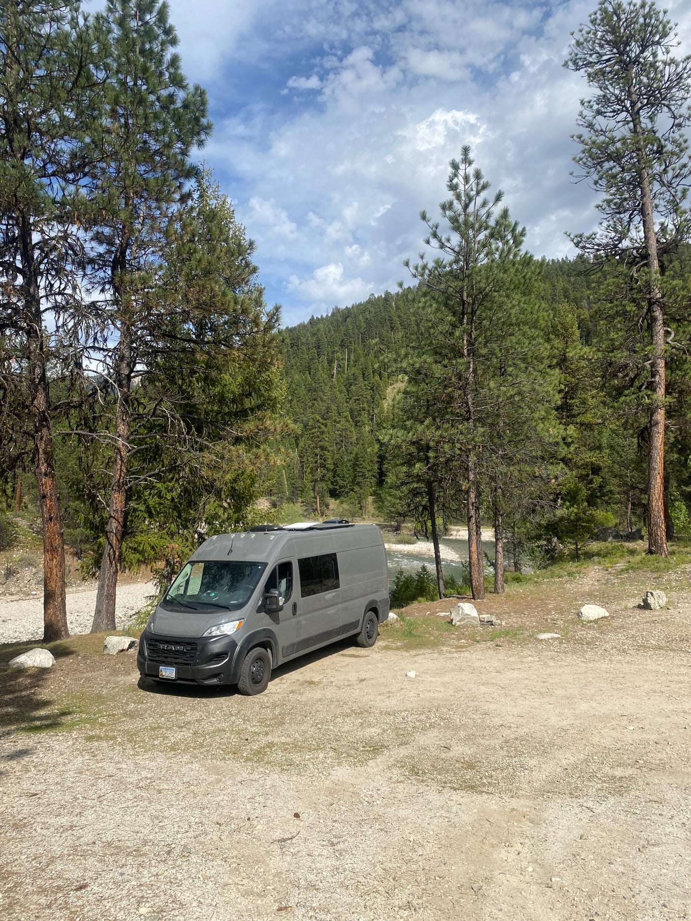 Gray camper van RV rental parked by a river in a mountainous forest setting under a cloudy blue sky.