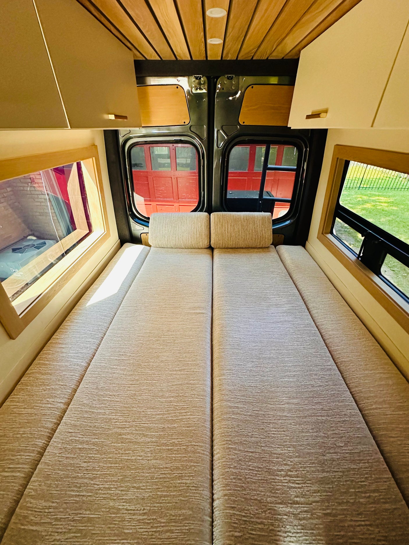 Cozy RV rental camper bed with wooden ceiling, overhead cabinets, and large side windows for scenic views.