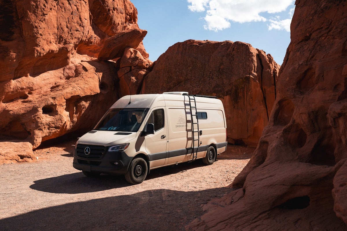 A beige Mercedes Sprinter **rv rental** campervan is parked on a dirt road amidst towering red rock formations, perfect for desert adventure.