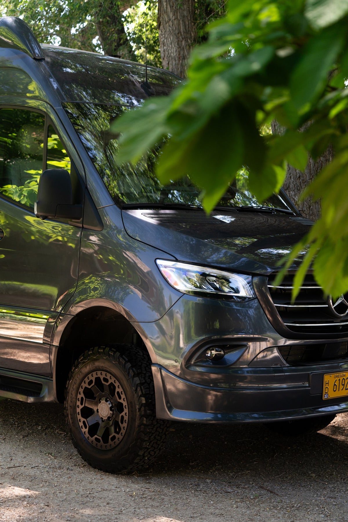 Dark gray Mercedes Sprinter camper van, an RV rental, with off-road tires, parked outdoors beneath leafy trees. — photo 18