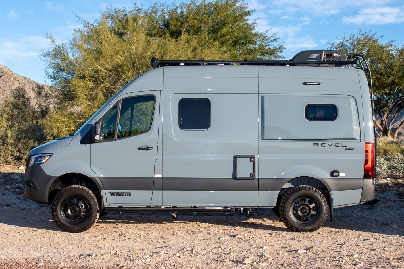 Side view of a gray Winnebago Revel 4x4 camper van parked on a dirt road. Ideal for an RV rental adventure.