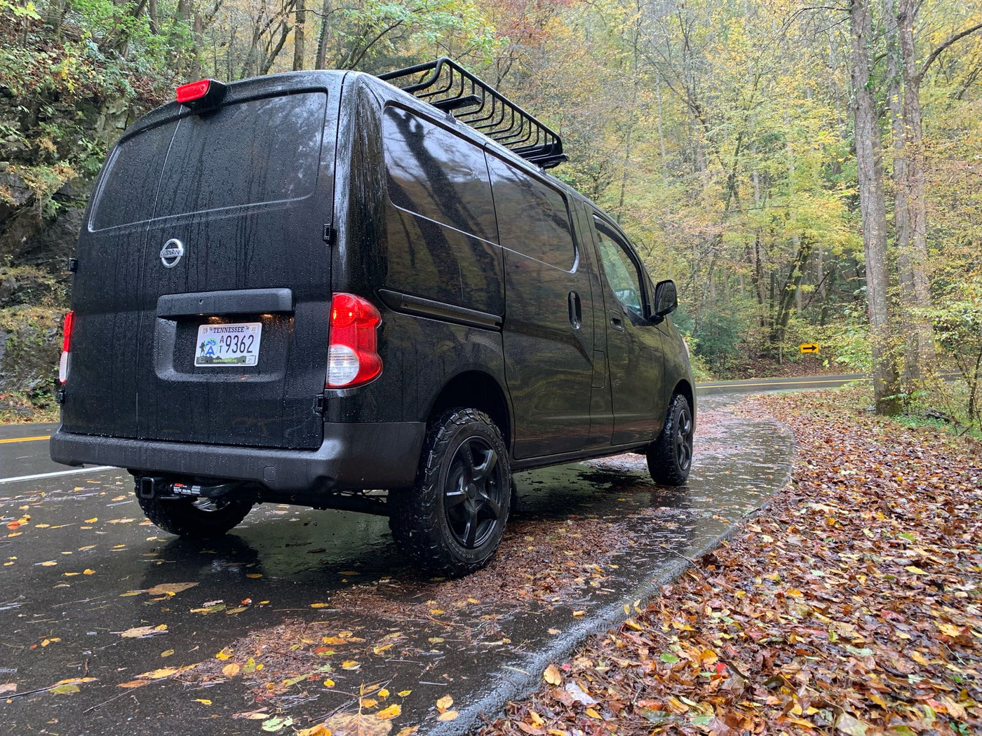 Black Nissan van, ideal for an RV rental road trip, parked on a wet road with autumn leaves & forest. — photo 9
