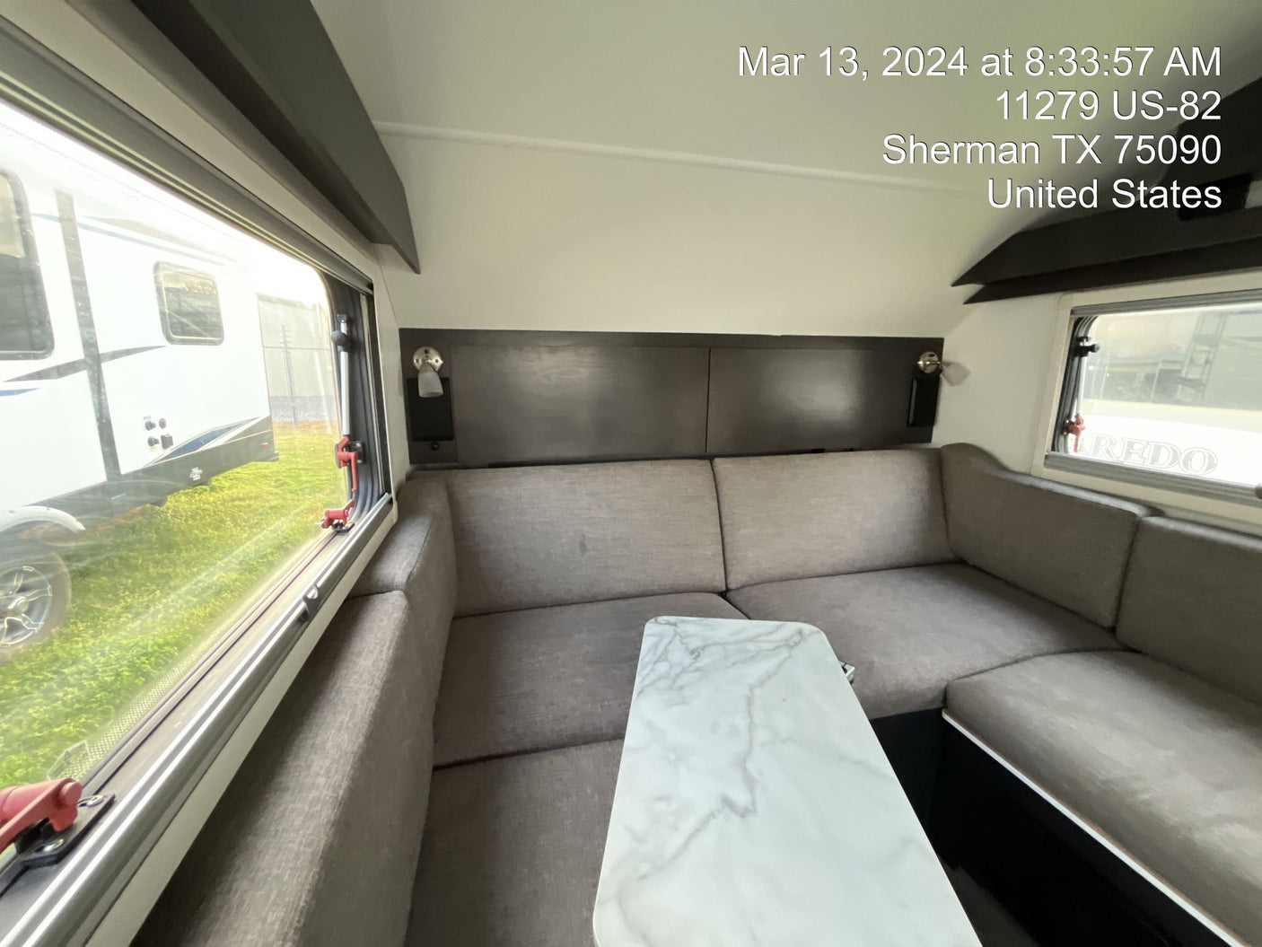 Spacious RV rental interior with a gray sectional couch and marble-look table; bright windows show an outside trailer.