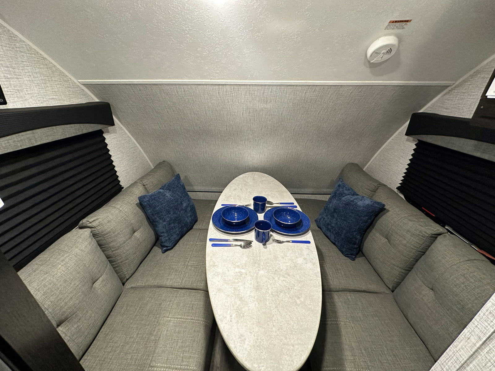 Inside an RV rental: a cozy U-shaped dinette with grey cushions, a white table set with blue dishes, and black window blinds.
