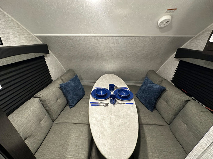 Inside an RV rental: a cozy U-shaped dinette with grey cushions, a white table set with blue dishes, and black window blinds.