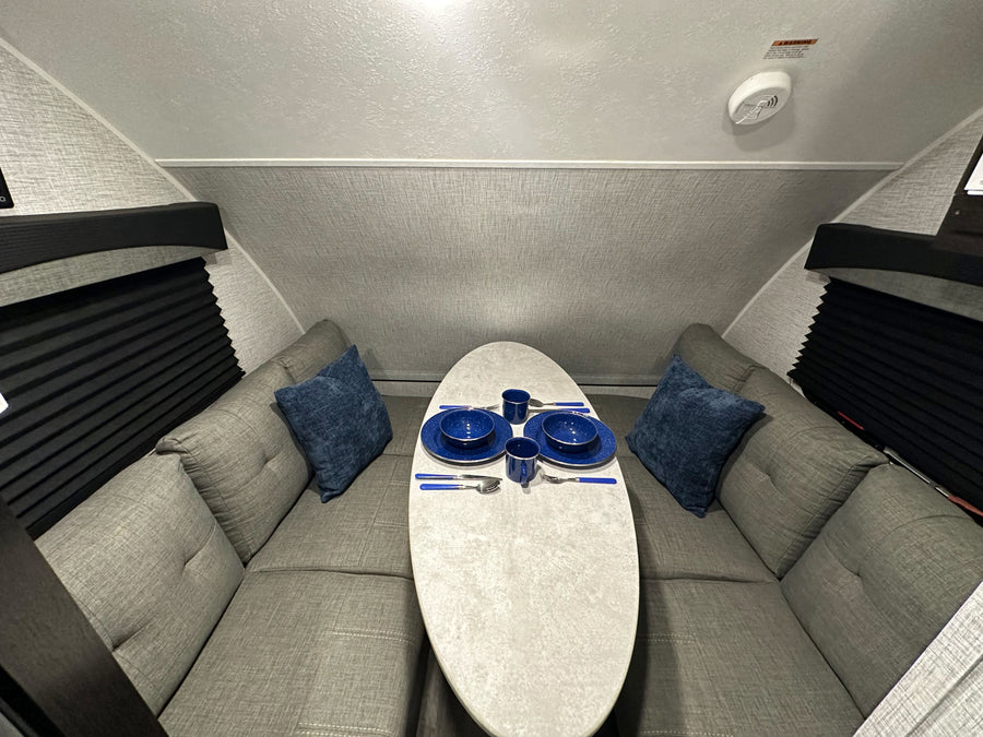 Inside an RV rental: a cozy U-shaped dinette with grey cushions, a white table set with blue dishes, and black window blinds.