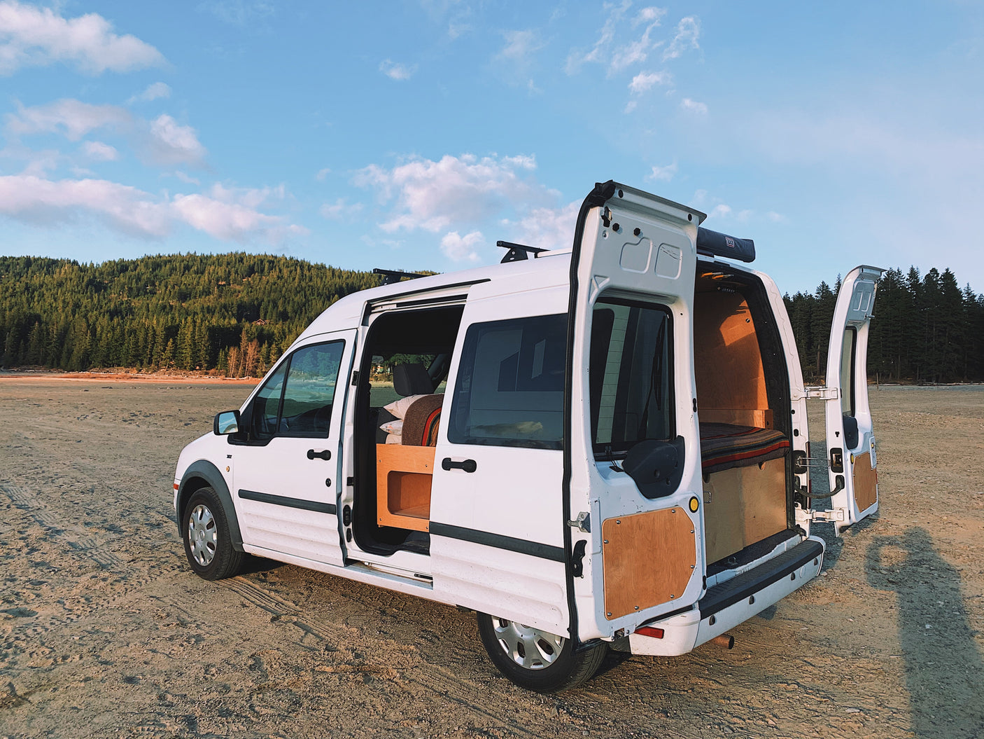 White camper van (RV rental) with doors open on a sandy shore. Inside, a cozy, custom-built bed. Scenic forest view.