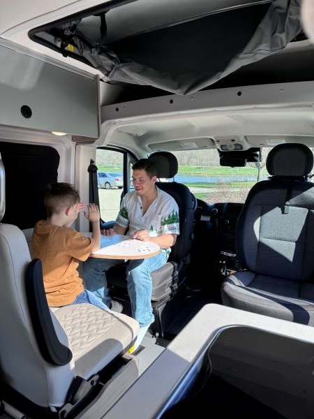 and table with a book & mug. Perfect for your next adventure!|Experience nature from your RV rental! Child gazing at a sunlit forest view from the cozy bed. Ideal family adventure.|RV rental kitchen with open overhead cabinets showing coffee pods