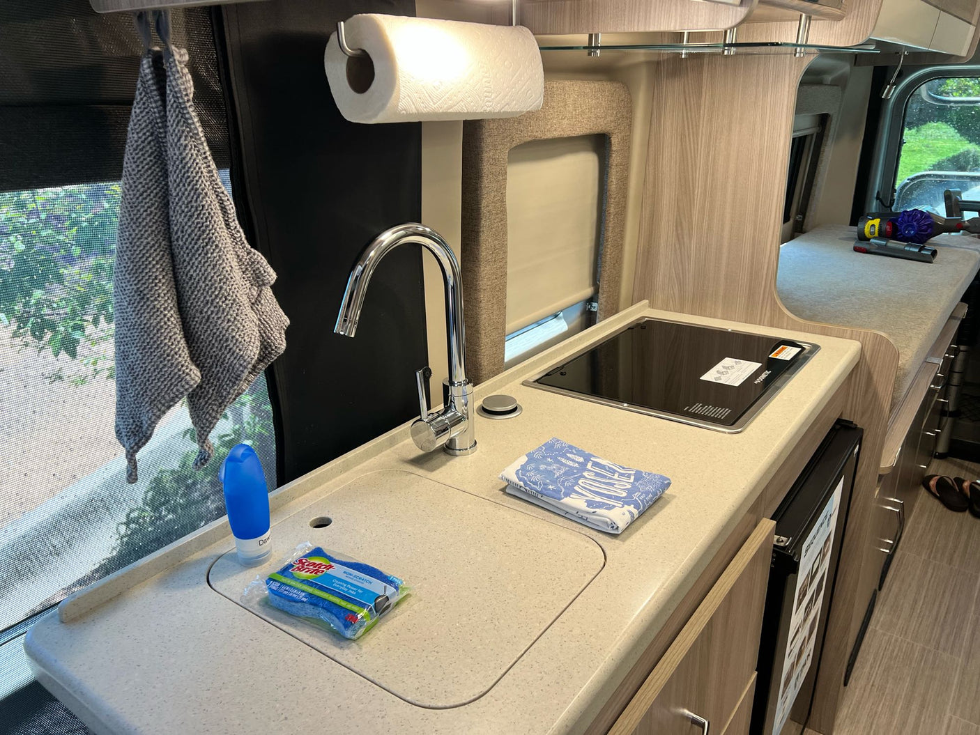 An RV rental's functional kitchen featuring a sink, cooktop, paper towels, and cleaning supplies. Ideal for on-the-go meals.