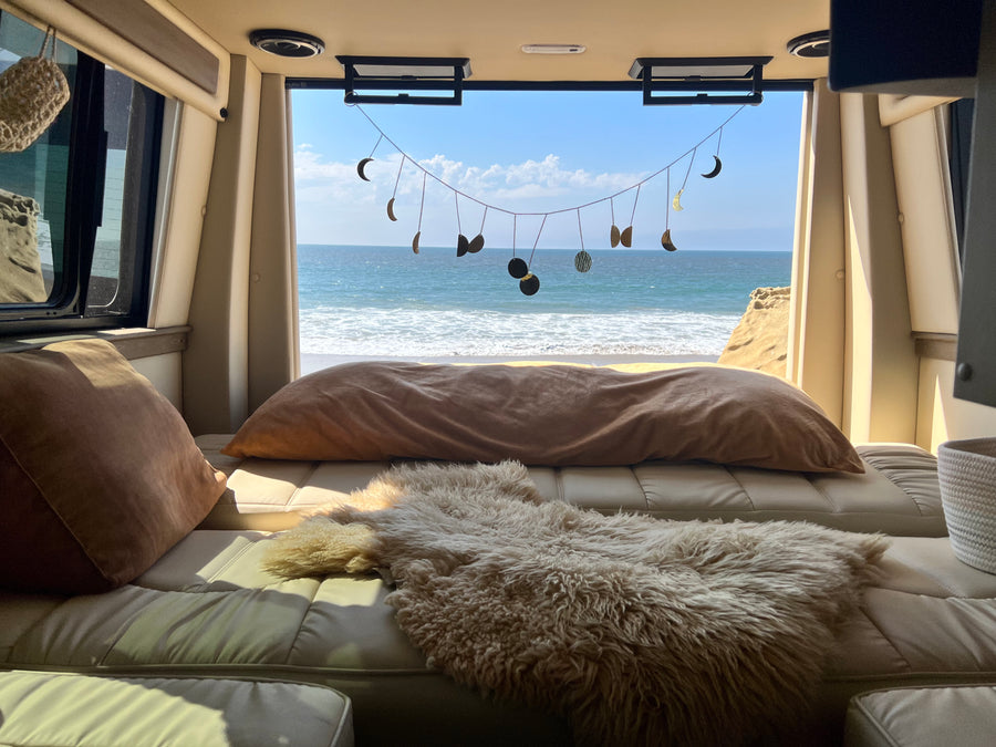 Relaxing RV rental interior with a cozy bed, pillows, and sheepskin rug overlooking a stunning ocean view and beach.