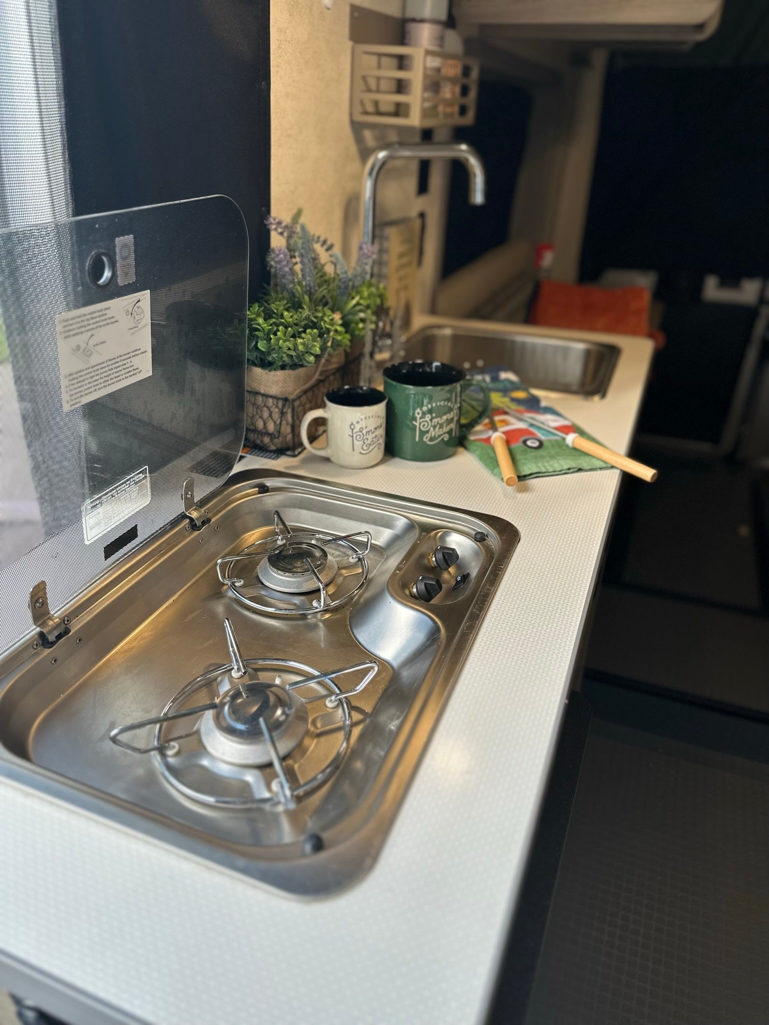 Compact RV rental kitchen featuring a two-burner stove, sink, and countertop with accessories. Ideal for cooking on the road.