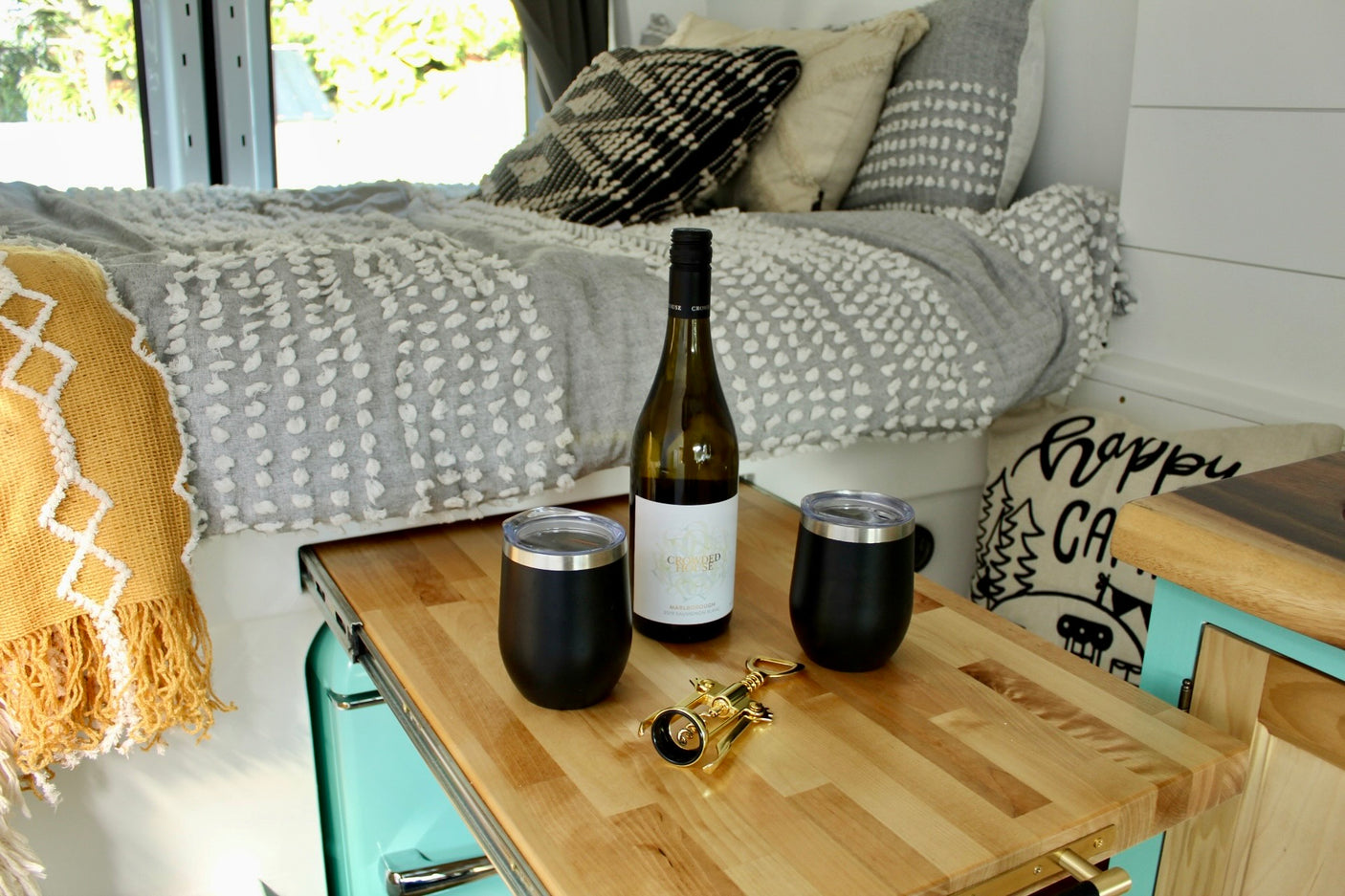 RV rental glamping: wine, glasses, corkscrew on wooden counter. Plush bed and throw blankets create a cozy camper van interior.