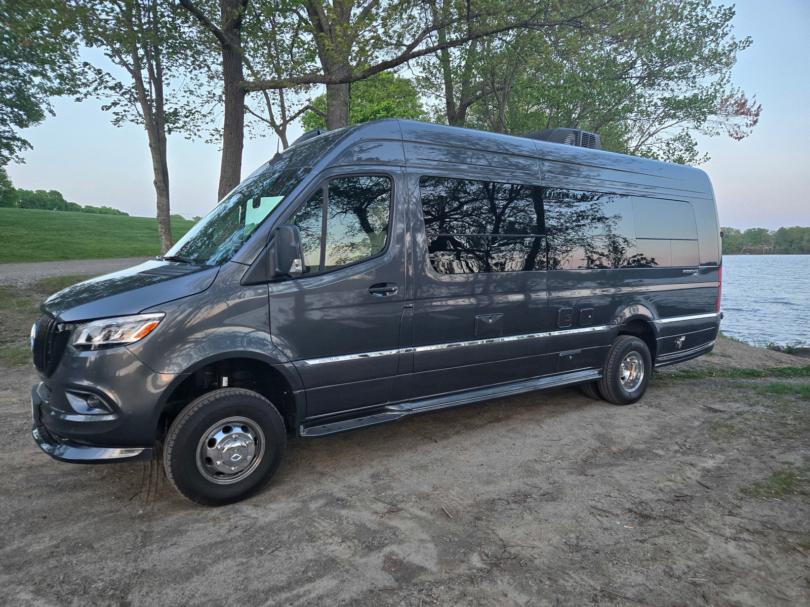 Dark gray Airstream Interstate AWD camper van (RV rental) parked by a lake, surrounded by trees.