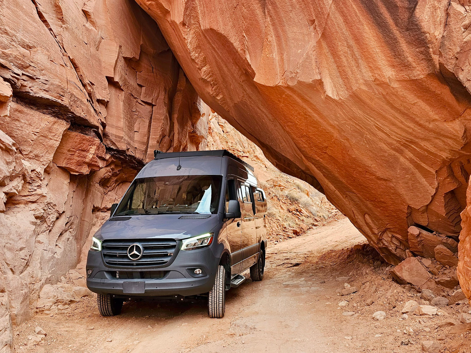 This grey camper van, perfect for rv rental adventures, navigates a rugged dirt path through a dramatic red rock canyon.