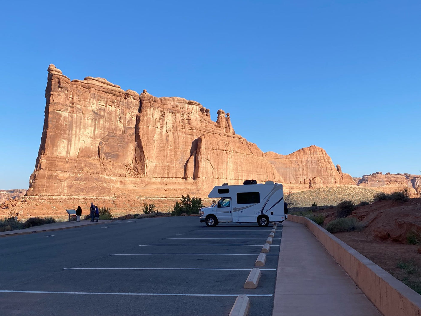 An RV rental sits in a sunlit parking lot, showcasing majestic red rock formations in a stunning desert national park.