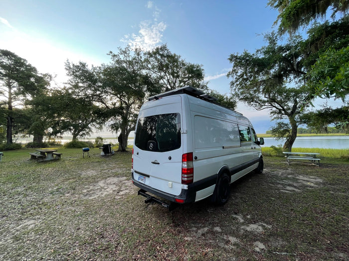 is parked on a dirt mountain road. Perfect for adventure!|White Sprinter van RV rental parked amidst lush trees with Spanish moss. Perfect for nature adventures & camping.|White Sprinter RV rental parked at a scenic waterfront campsite with trees. Ideal for outdoor adventures & van life.|White Mercedes-Benz Sprinter van RV rental parked on a dirt path with trees. Kayaks and grill visible in background.|White Sprinter van RV rental parked on a grassy bank by a wide blue lake under a clear sky. Ready for ad