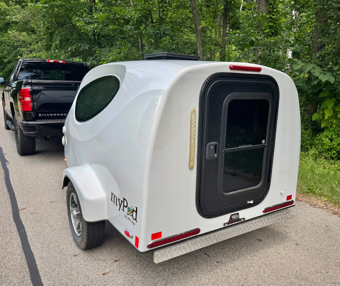 Explore nature with this myPod teardrop RV rental, hitched to a black pickup truck, amidst green trees.