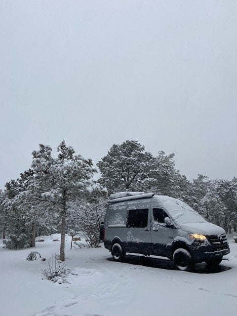 Snow-covered gray Sprinter RV rental van nestled in a serene winter forest. A peaceful travel scene.