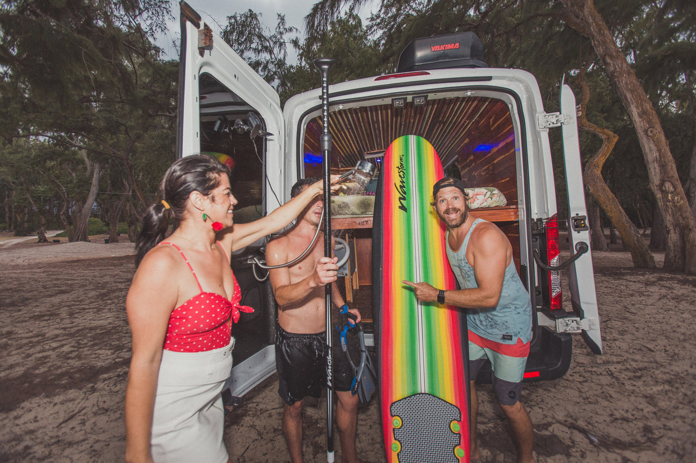 Three friends at their RV rental, with one showering by the van and another pointing at a colorful surfboard, ready for a beach day.