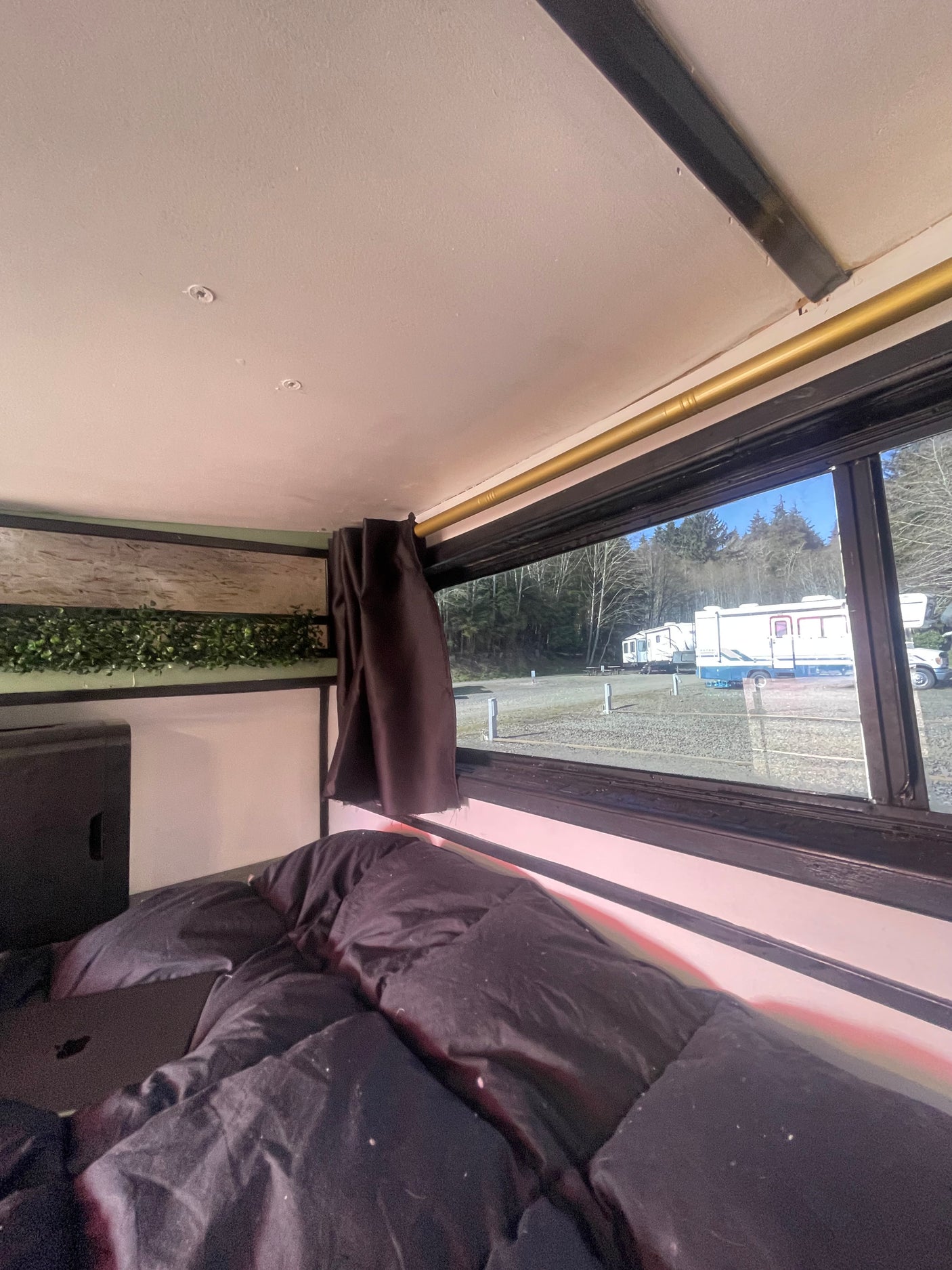 Cozy interior of an RV rental with a black comforter, looking out to a campground with other RVs and trees.